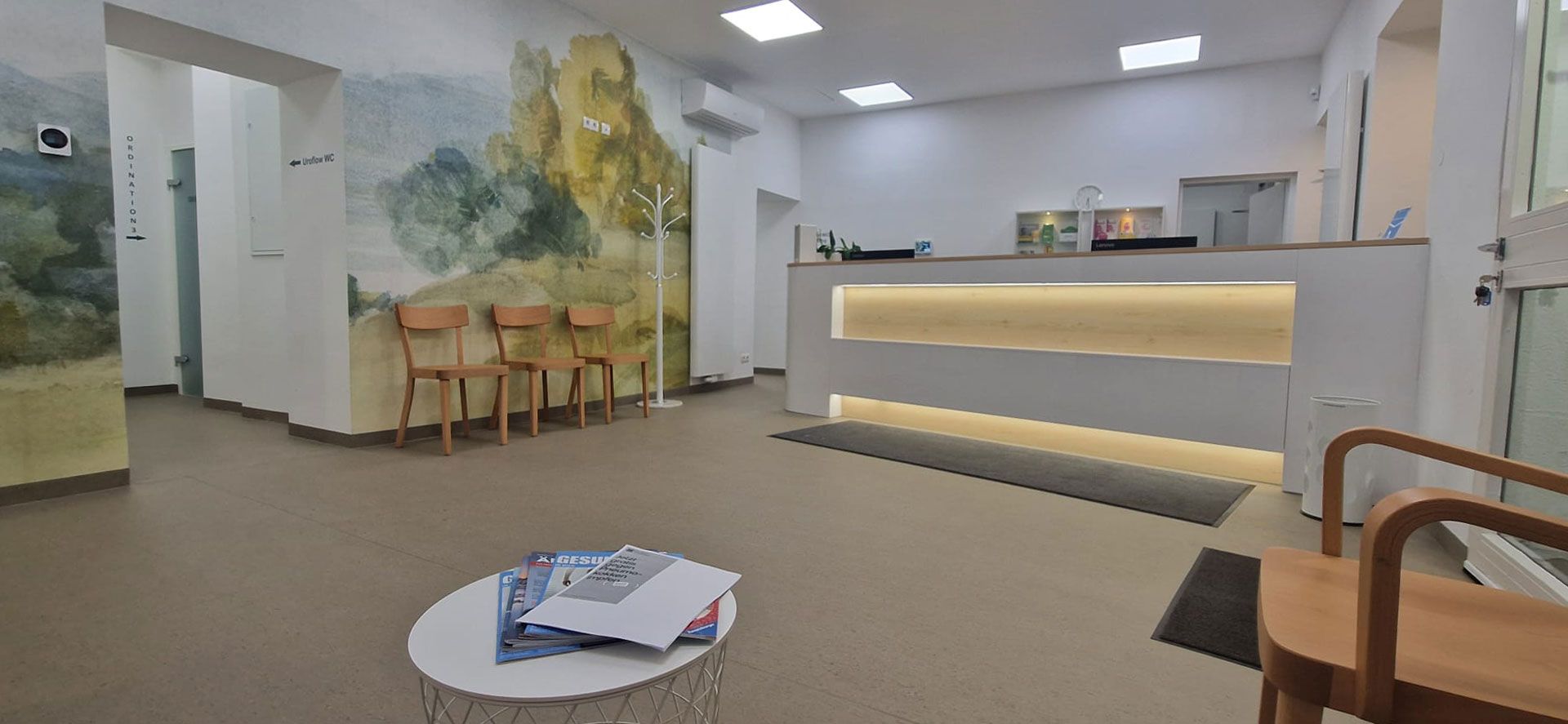 Wartebereich mit rundem Tisch, mehreren Holzstühlen und beleuchtetem Empfangstresen in einem hellen Raum