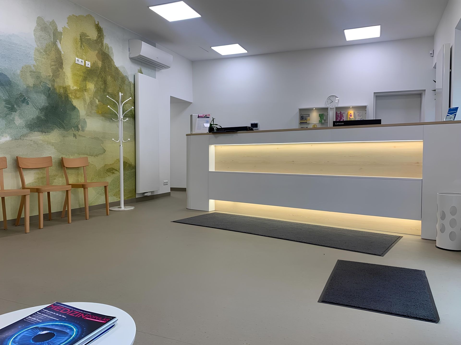 Heller Empfangsbereich mit beleuchtetem Tresen, mehreren Holzstühlen vor einer Wand mit grünem Muster und Garderobenständer